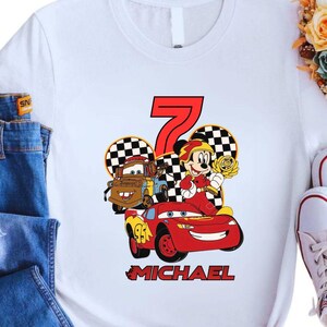 May include: White t-shirt featuring a colorful graphic of Lightning McQueen, Mater, and Mickey Mouse celebrating a seventh birthday. The design includes the number 7, checkered flags, and the name "MICHAEL" in red.