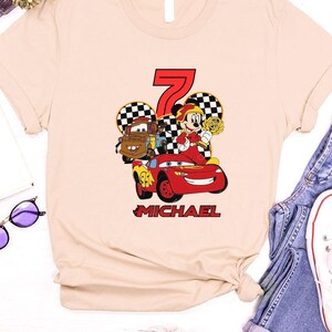May include: A light beige t-shirt with a vibrant graphic of the number 7, race cars, and a cartoon character. The name "MICHAEL" is printed in red below the graphic. This t-shirt is designed for casual wear.