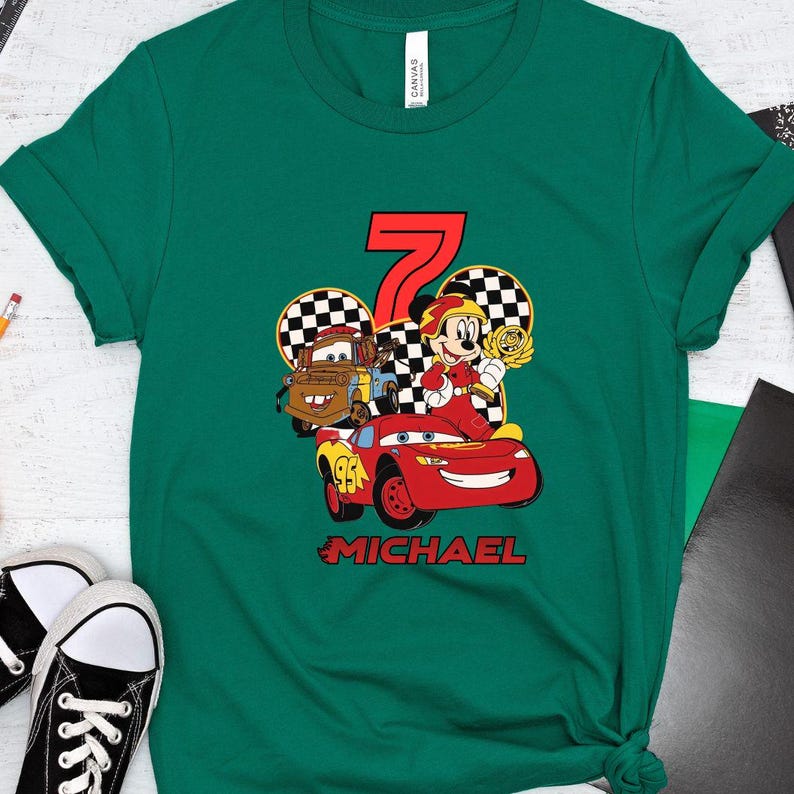 May include: Green t-shirt with a vibrant graphic of Disney's Cars characters, including Lightning McQueen, Mater, and Mickey Mouse. The number 7 is in red, with "MICHAEL" below. A fun birthday shirt for a child.