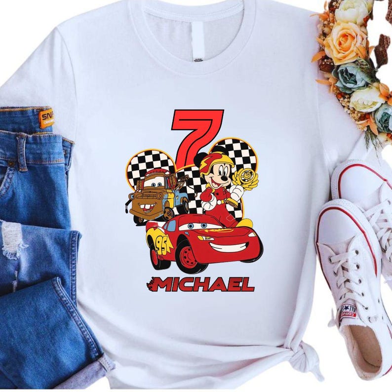 May include: White t-shirt featuring a colorful graphic of Lightning McQueen, Mater, and Mickey Mouse celebrating a seventh birthday. The design includes the number 7, checkered flags, and the name "MICHAEL" in red.