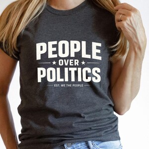 May include: Dark gray t-shirt with the words "PEOPLE OVER POLITICS" in white block letters. The text includes two stars and the phrase "EST. WE THE PEOPLE" below the main text. The shirt is a crew neck style.