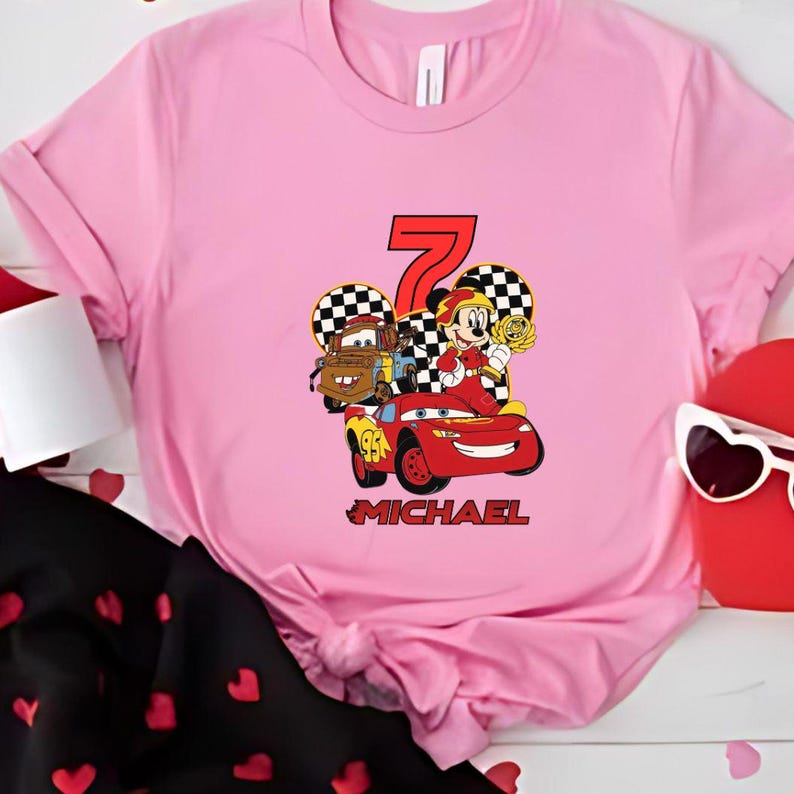 May include: A pink t-shirt with a graphic of Disney's Cars characters, including Lightning McQueen, Mater, and Mickey Mouse. The number 7 is in red, with the name "MICHAEL" below. The shirt has a crew neck and is made of a soft material.