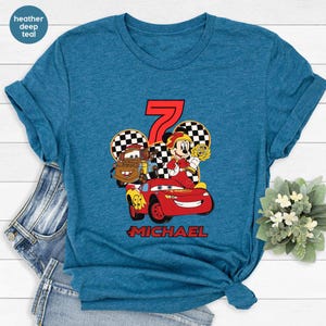 May include: Teal t-shirt with a vibrant graphic of a red race car, a cartoon character, and the number 7. The name "MICHAEL" is printed below the graphic. The shirt is made of a soft material.