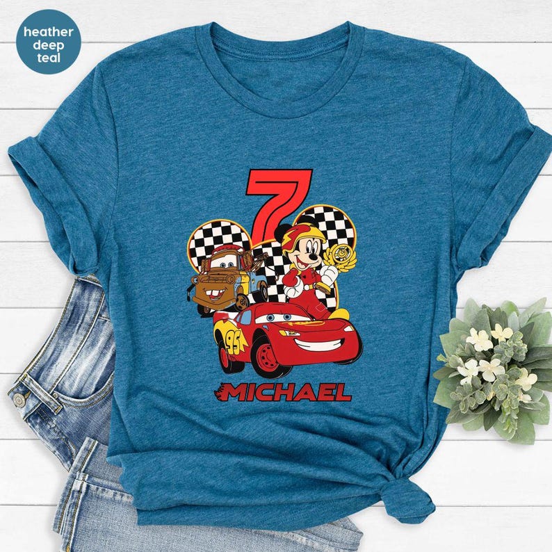 May include: Teal t-shirt with a vibrant graphic of a red race car, a cartoon character, and the number 7. The name "MICHAEL" is printed below the graphic. The shirt is made of a soft material.