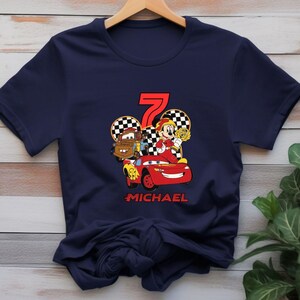 May include: Navy blue t-shirt with a graphic of a red race car, Mater, and Mickey Mouse holding a trophy. The number 7 is in red, with the name "MICHAEL" below the car. The design is set against a checkered background.