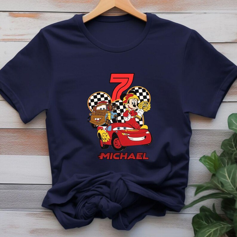 May include: Navy blue t-shirt with a graphic of a red race car, Mater, and Mickey Mouse holding a trophy. The number 7 is in red, with the name "MICHAEL" below the car. The design is set against a checkered background.