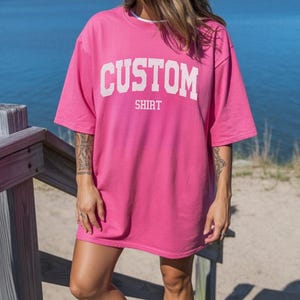 May include: A neon pink oversized t-shirt with the word "CUSTOM" printed in white block letters. The word "SHIRT" is printed below "CUSTOM". The shirt is worn with white sneakers and socks. The background includes a wooden staircase and a body of water.