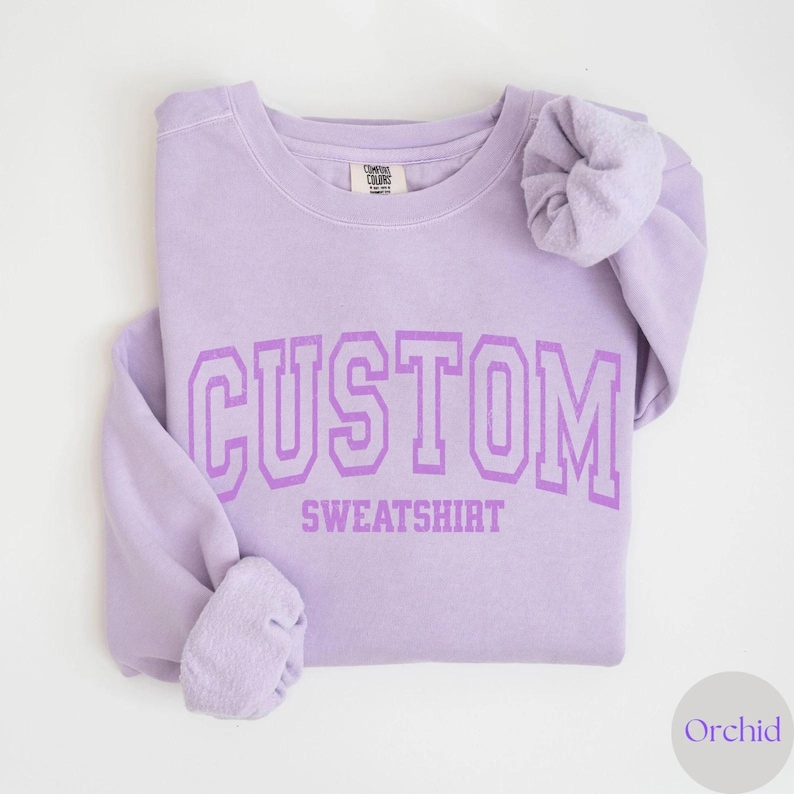 May include: A lilac sweatshirt featuring the word "CUSTOM" above "SWEATSHIRT" in a matching purple, collegiate-style font. The sleeves are folded. The sweatshirt is on a white background. The colour is labelled "Orchid".