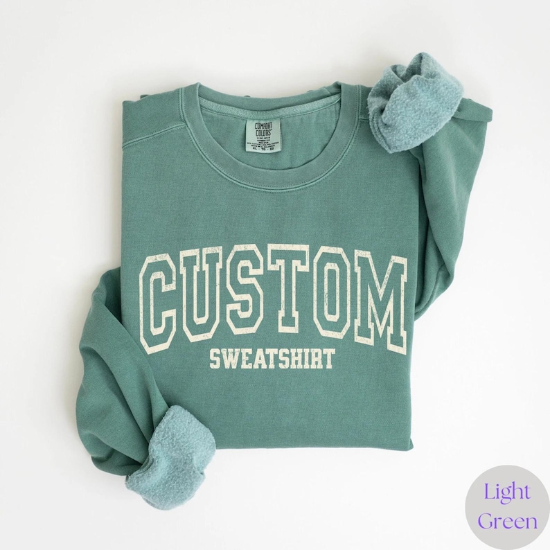 May include: A light green sweatshirt with the text "CUSTOM SWEATSHIRT" printed in a white, collegiate-style font. The sweatshirt is folded, with the sleeves and collar visible. The fabric appears soft and comfortable.