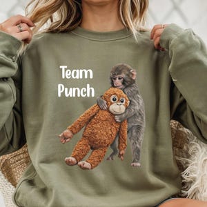May include: An olive green sweatshirt with the text "Team Punch" in white. The sweatshirt features a graphic of two monkeys hugging. One monkey is brown, and the other is gray.