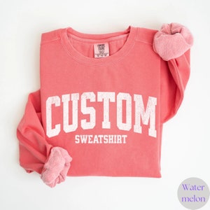 May include: A coral-coloured sweatshirt with the word "CUSTOM" in large, white, block letters above "SWEATSHIRT". The sleeves are folded, and a matching scrunchie is on the right sleeve. The tag reads "COMFORT COLORS".
