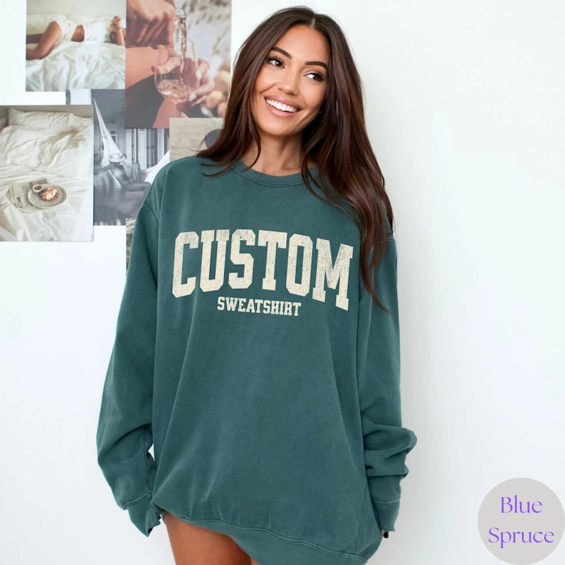 May include: Teal sweatshirt featuring the word "CUSTOM" in large, off-white, distressed lettering. The word "SWEATSHIRT" is printed below in a smaller font. The sweatshirt has a relaxed fit. The brand name "Blue Spruce" is in a circle at the bottom right.
