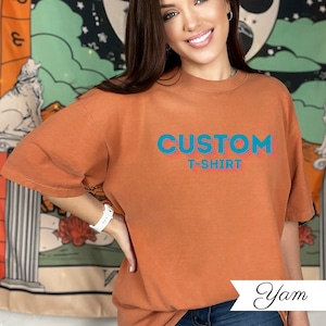 May include: An orange t-shirt with the words "CUSTOM T-SHIRT" in blue and red. The shirt is a solid colour and has short sleeves. The model is wearing a white watch.