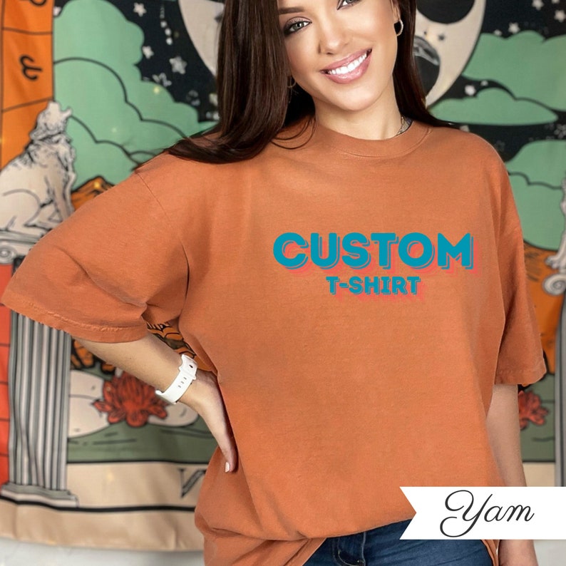 May include: An orange t-shirt with the words "CUSTOM T-SHIRT" in blue and red. The shirt is a solid colour and has short sleeves. The model is wearing a white watch.