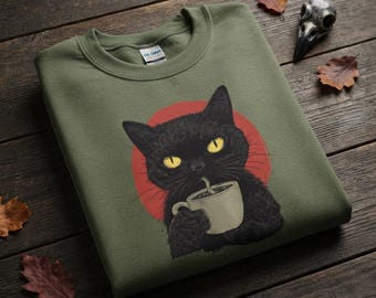 May include: An olive green crewneck sweatshirt featuring a graphic illustration of a black cat with bright yellow eyes holding a beige mug. The cat is positioned in front of a solid red circle. The inner collar tag displays the brand name Gildan.