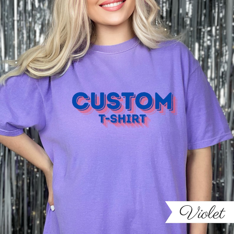 May include: A violet t-shirt featuring the text "CUSTOM T-SHIRT" in blue with a pink shadow. The shirt has a casual fit and is a solid colour. The word "Violet" is written in cursive in the bottom right corner of the image.