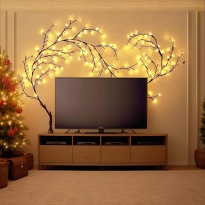 May include: A living room scene featuring a large flat-screen TV mounted above a wooden media console. Decorative branches with warm white fairy lights adorn the wall, creating a cozy ambiance. A decorated Christmas tree is visible on the left.