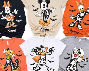May include: A selection of Halloween t-shirts featuring Disney characters dressed as skeletons. The shirts are in black, beige, orange, white, and grey. Each shirt displays a character design with the word "Name" printed underneath.