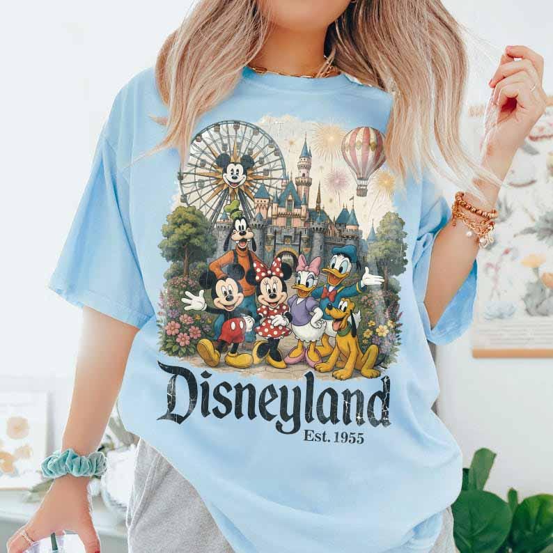 May include: A light blue t-shirt featuring a graphic print of Mickey Mouse, Minnie Mouse, Goofy, Donald Duck, Daisy Duck, and Pluto in front of a castle, a Ferris wheel, and a hot air balloon. The text reads Disneyland Est. 1955 in black lettering.
