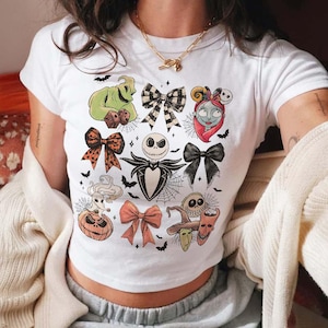 May include: White t-shirt featuring colorful illustrations of characters and bows from The Nightmare Before Christmas. The design includes Jack Skellington, Sally, Oogie Boogie, and other iconic elements, perfect for fans of the movie.