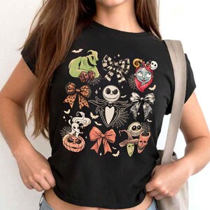 May include: Black t-shirt featuring a collage of characters and bow designs from The Nightmare Before Christmas. The design includes Jack Skellington, Sally, Oogie Boogie, and other Halloween-themed elements. The shirt is a crew neck.