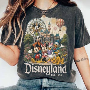May include: A charcoal grey t-shirt featuring a graphic print of Mickey Mouse, Minnie Mouse, Goofy, Donald Duck, Daisy Duck, and Pluto in front of a castle, a Ferris wheel, and a hot air balloon. The text reads Disneyland Est. 1955.