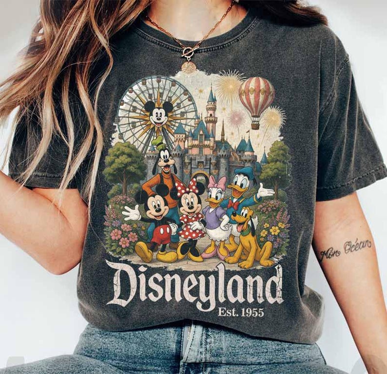 May include: A charcoal grey t-shirt featuring a graphic print of Mickey Mouse, Minnie Mouse, Goofy, Donald Duck, Daisy Duck, and Pluto in front of a castle, a Ferris wheel, and a hot air balloon. The text reads Disneyland Est. 1955.