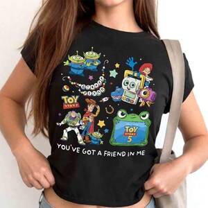 K&ouml;nnte beinhalten: Ein schwarzes T-Shirt mit einem farbenfrohen Grafikdruck von Toy Story-Figuren, darunter Buzz Lightyear, Woody und die Aliens. Das Design enth&auml;lt den Text Disney Pixar, Toy Story, Toy Story 5 und den Schriftzug You've Got A Friend In Me.