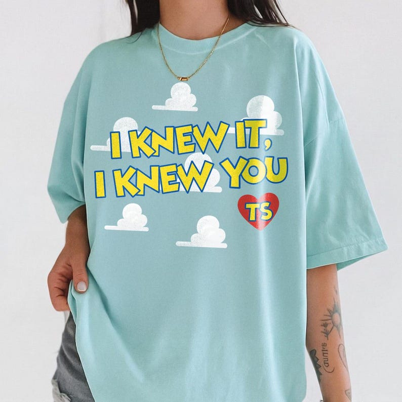 Może przedstawiać: Jasnoniebieski t-shirt z kr&oacute;tkim rękawem z nadrukiem białych chmur oraz ż&oacute;łtym napisem I KNEW IT, I KNEW YOU i czerwonym sercem z literami TS. Koszulka posiada okrągły dekolt oraz luźny, swobodny kr&oacute;j.