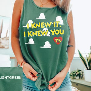 May include: A light green sleeveless tank top featuring a graphic print of white clouds and the yellow text I KNEW IT, I KNEW YOU with a red heart containing the letters TS. The garment has a round neckline and a relaxed fit design.