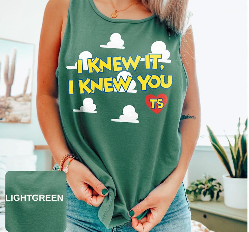 May include: A light green sleeveless tank top featuring a graphic print of white clouds and the yellow text I KNEW IT, I KNEW YOU with a red heart containing the letters TS. The garment has a round neckline and a relaxed fit design.