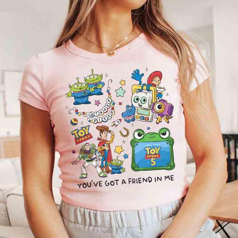 K&ouml;nnte beinhalten: Ein hellrosa T-Shirt mit kurzen &Auml;rmeln und einem farbenfrohen Grafikdruck von Toy Story-Figuren, darunter Woody, Buzz Lightyear und Aliens. Das Design enth&auml;lt den Text Disney Pixar, Toy Story, Toy Story 5 und You've Got A Friend In Me.