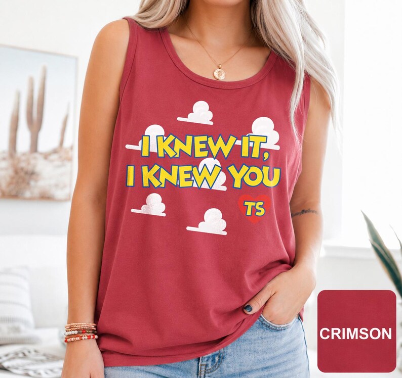 May include: A crimson sleeveless tank top featuring a graphic print of white clouds and the yellow text "I KNEW IT, I KNEW YOU" with a red heart containing the letters "TS" positioned below the text on the front of the garment.