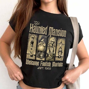 May include: Black t-shirt featuring a graphic from The Haunted Mansion. The design includes the text "The Haunted Mansion" above four character illustrations, with "Welcome Foolish Mortals" and "EST. 1969" below. The design is in gold.
