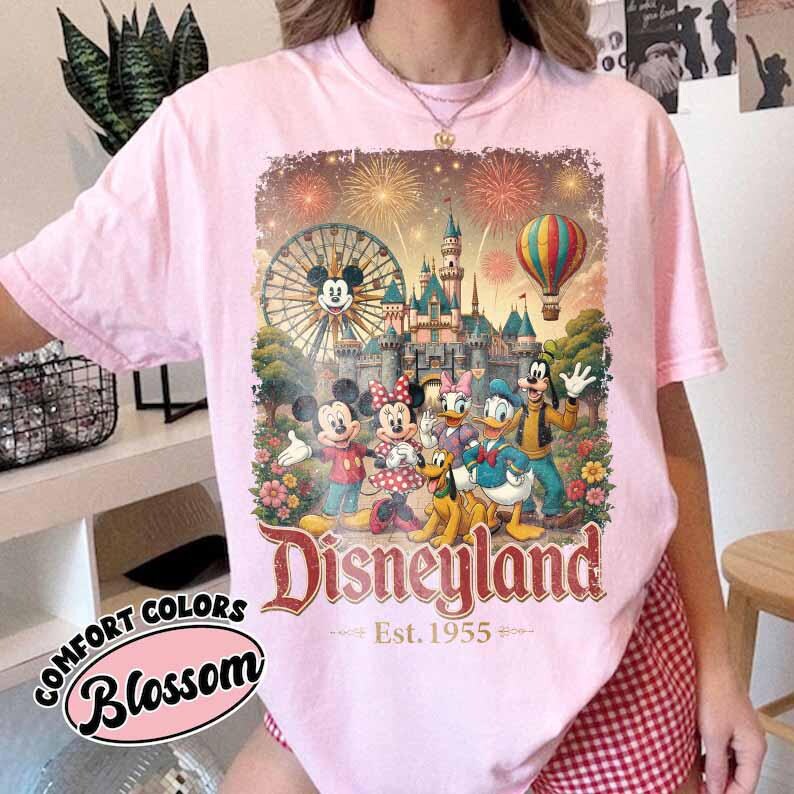 May include: A light pink short-sleeve t-shirt featuring a graphic print of Mickey Mouse, Minnie Mouse, Pluto, Donald Duck, Daisy Duck, and Goofy in front of a castle, a Ferris wheel, and a hot air balloon with fireworks. The text reads Disneyland Est. 1955.