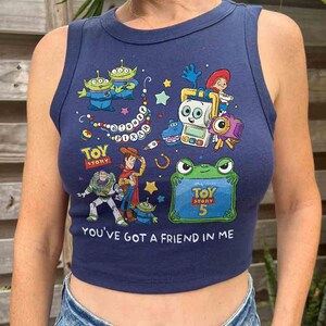 K&ouml;nnte beinhalten: Ein &auml;rmelloses, dunkelblaues bauchfreies Oberteil mit einem bunten Toy Story-Grafikdruck. Das Design zeigt Figuren wie Woody, Buzz Lightyear und Aliens sowie den Text Disney Pixar, Toy Story 5 und You've Got A Friend In Me.
