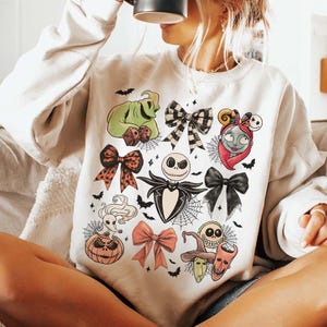 May include: Cream-coloured sweatshirt with a colourful graphic of characters and bows from "The Nightmare Before Christmas." The design includes Jack Skellington, Sally, Oogie Boogie, and other iconic elements, with bats and spiderwebs.
