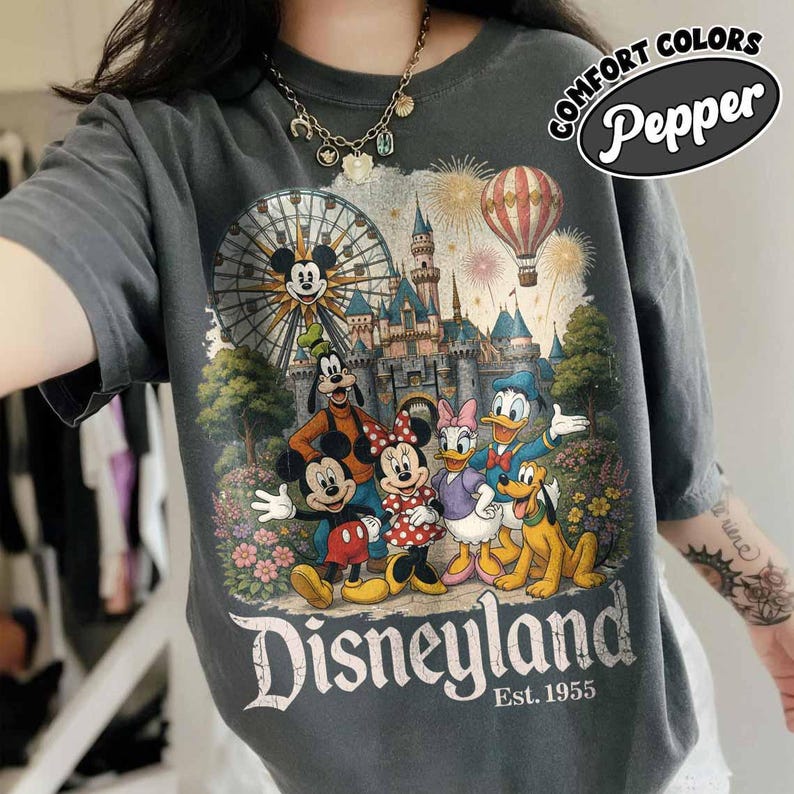 May include: A charcoal grey t-shirt featuring a graphic print of Mickey Mouse, Minnie Mouse, Goofy, Donald Duck, Daisy Duck, and Pluto in front of a castle, a Ferris wheel, and a hot air balloon. The text reads Disneyland Est. 1955 and Comfort Colors Pepper.