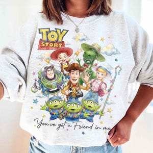 May include: A light gray sweatshirt featuring the words "Toy Story" above a colorful graphic of the movie's characters, including Woody, Buzz Lightyear, and the Little Green Men. The phrase "You've got a Friend in me" is written below the graphic.