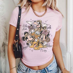 Pu&ograve; includere: T-shirt rosa chiaro con grafica dei personaggi Disney Topolino, Minnie, Paperino, Paperina, Pippo e Pluto. I personaggi indossano cappelli da safari. La maglietta ha il testo "Mickey Mouse" e "Minnie Mouse" in corsivo.
