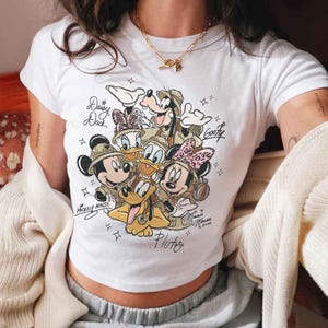 Pu&ograve; includere: T-shirt bianca con una grafica dei personaggi Disney Topolino, Minnie, Paperino, Paperina, Pippo e Pluto in tenute da safari. I nomi dei personaggi sono scritti sulla maglietta. La maglietta ha un girocollo e maniche corte.