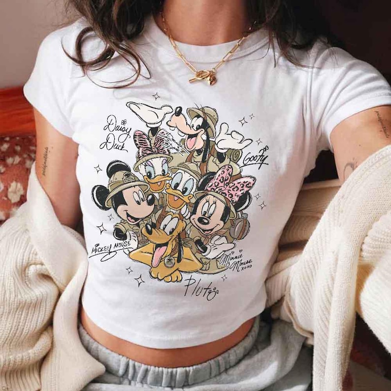 Pu&ograve; includere: T-shirt bianca con una grafica dei personaggi Disney Topolino, Minnie, Paperino, Paperina, Pippo e Pluto in tenute da safari. I nomi dei personaggi sono scritti sulla maglietta. La maglietta ha un girocollo e maniche corte.