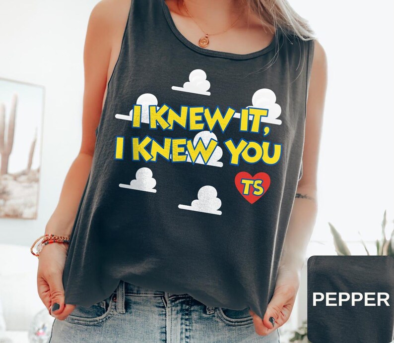 May include: A charcoal gray sleeveless tank top featuring a graphic print of white clouds and yellow text that reads I KNEW IT, I KNEW YOU with a red heart containing the letters TS. The garment has a round neckline and a relaxed fit.