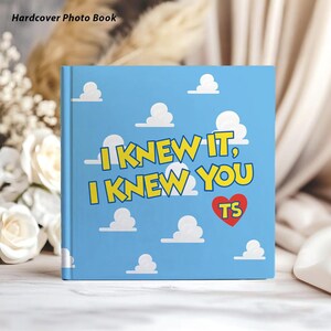 May include: A square hardcover photo book with a light blue cover featuring a repeating pattern of white clouds. The text "I KNEW IT, I KNEW YOU" is printed in yellow with a red heart containing the letters "TS" in the center of the front cover.