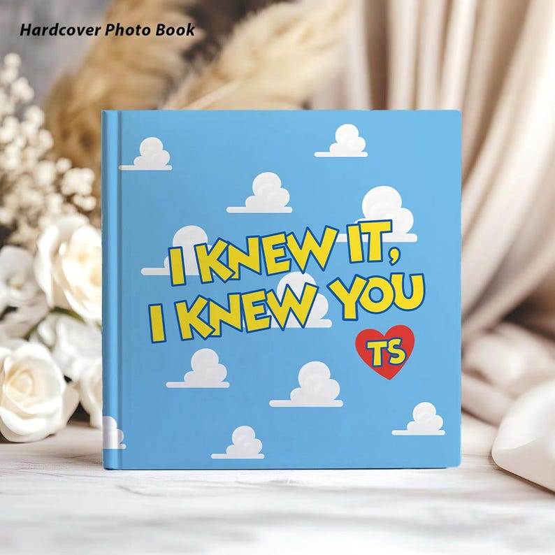 May include: A square hardcover photo book with a light blue cover featuring a repeating pattern of white clouds. The text "I KNEW IT, I KNEW YOU" is printed in yellow with a red heart containing the letters "TS" in the center of the front cover.