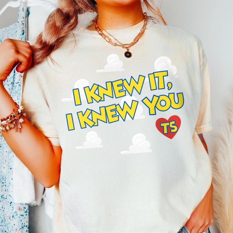 Może przedstawiać: Kremowy t-shirt z ż&oacute;łtym napisem I KNEW IT, I KNEW YOU oraz czerwonym sercem zawierającym litery TS. Wz&oacute;r zawiera białe grafiki chmur rozmieszczone na przodzie, nadrukowane na odzieży z kr&oacute;tkim rękawem i okrągłym dekoltem.