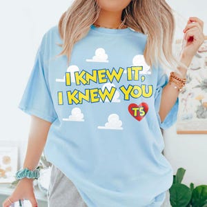 Może przedstawiać: Jasnoniebieski t-shirt z grafiką białych chmur i ż&oacute;łtym napisem I KNEW IT, I KNEW YOU. Pod tekstem znajduje się czerwone serce z ż&oacute;łtymi literami TS w środku. Koszulka ma kr&oacute;tkie rękawy i okrągły dekolt.