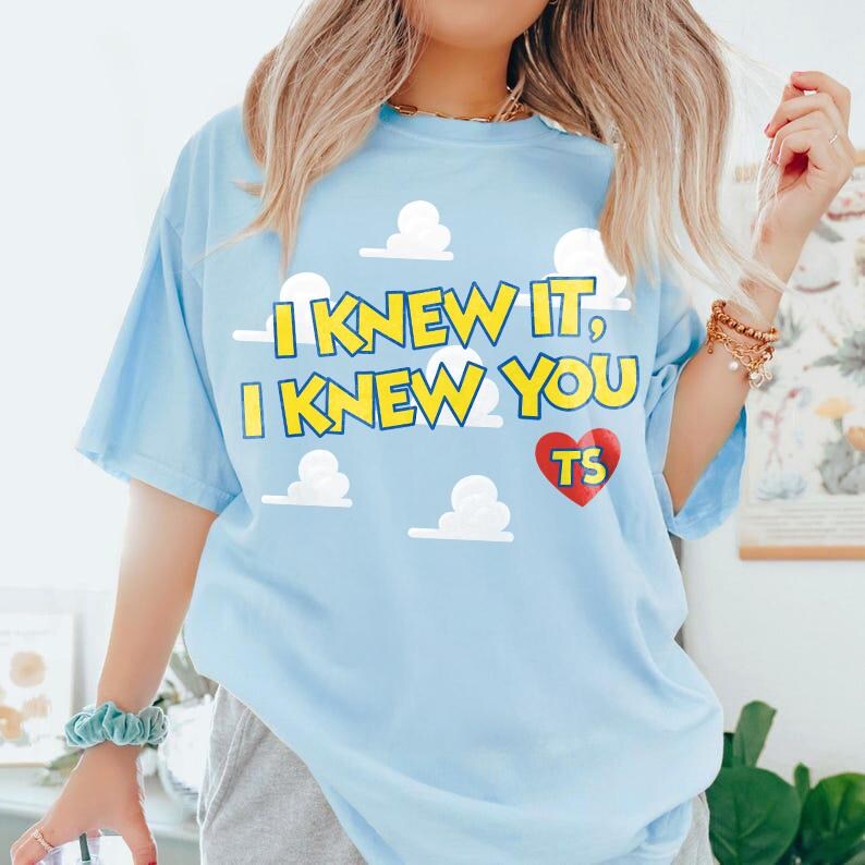 Może przedstawiać: Jasnoniebieski t-shirt z grafiką białych chmur i ż&oacute;łtym napisem I KNEW IT, I KNEW YOU. Pod tekstem znajduje się czerwone serce z ż&oacute;łtymi literami TS w środku. Koszulka ma kr&oacute;tkie rękawy i okrągły dekolt.
