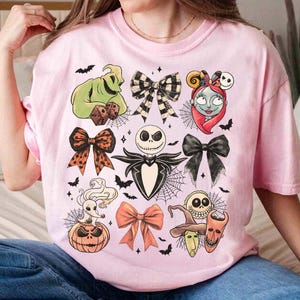 May include: A pale pink t-shirt with a Halloween-themed design. The graphic features characters and bow illustrations from "The Nightmare Before Christmas", along with bats and spiderwebs. The shirt is made of a soft material.