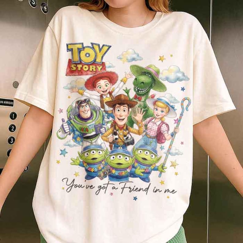 May include: Cream-colored t-shirt featuring a colorful graphic of Toy Story characters, including Woody, Buzz Lightyear, and the Little Green Men. The words "TOY STORY" are at the top, and the phrase "You've got a Friend in me" is at the bottom.